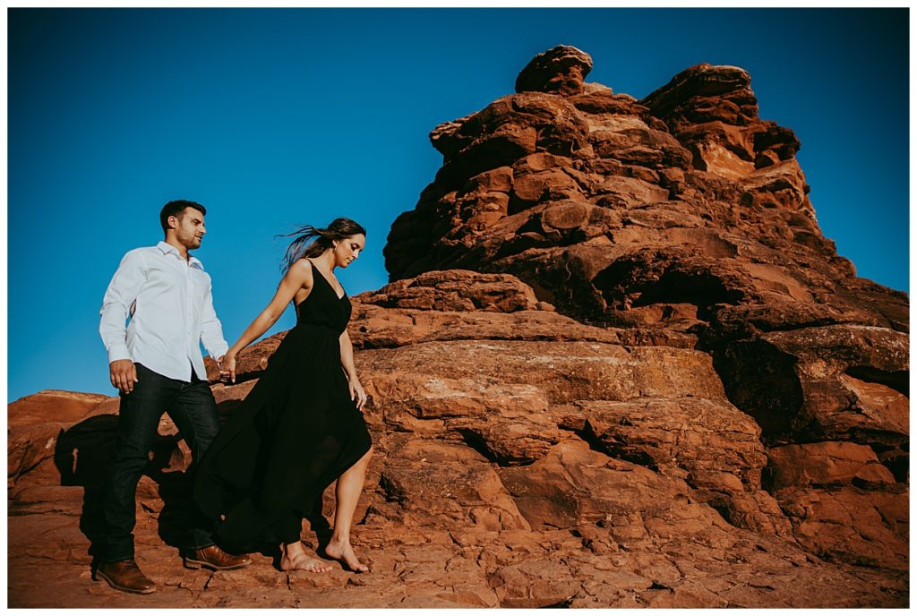 A man wearing a button down white shirt follows his fiance as she walks barefoot with a black cocktail dress during their Boynton Canyon Engagement photos