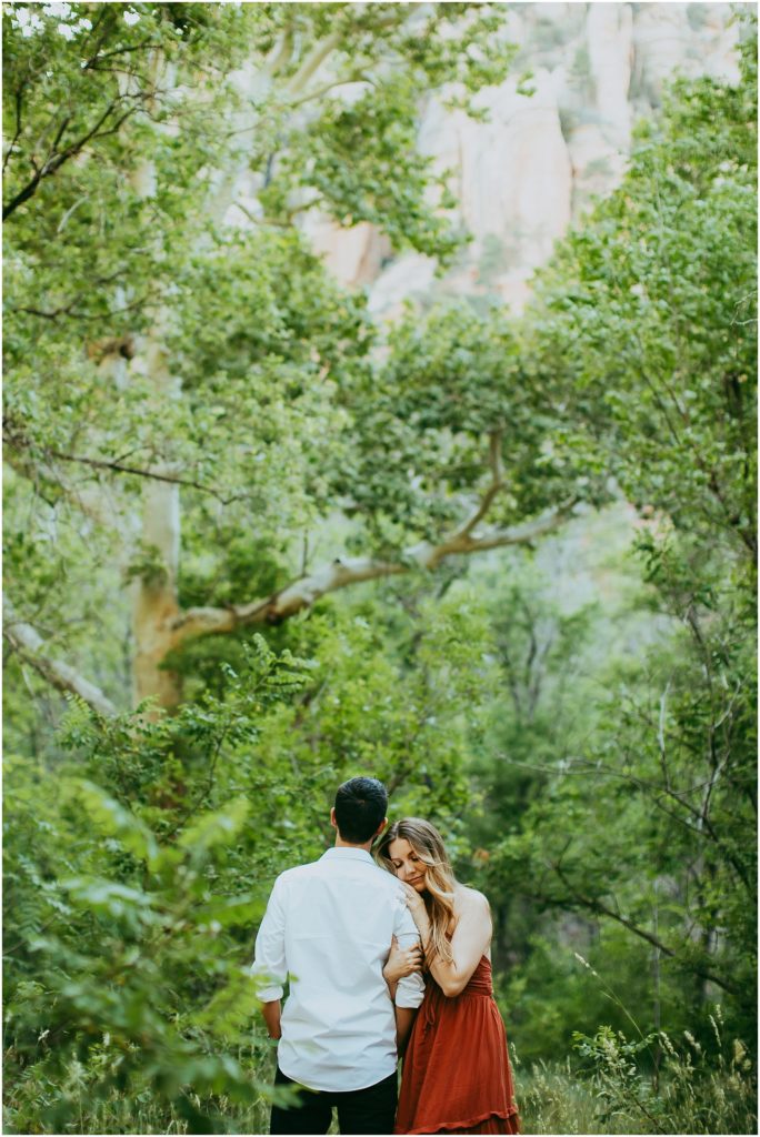 woman closes her eyes while leaning on man's shoulders among trees and grasses of oak creek canyon