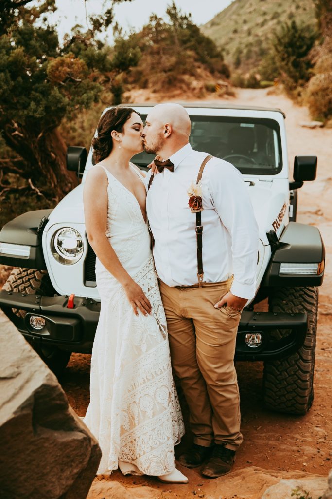 bride and groom kiss on the road to Merry Go Round Rock, posed in front of their white Rubicon Jeep.