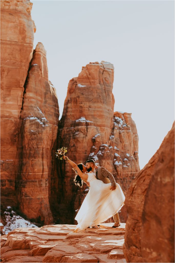 groom holds bride's leg to his hip as they kiss at Cathedral Rock in snowy Sedona