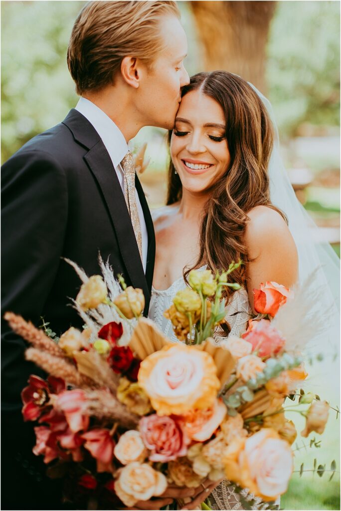 groom kisses bride's forehead with bouquet of beautiful flowers