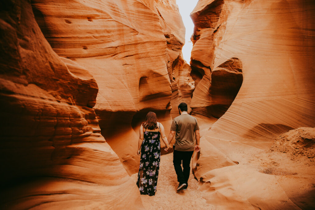 a man and woman in a dress hold hands and walk away from the camera as they walk through Secret Antelope Canyon.