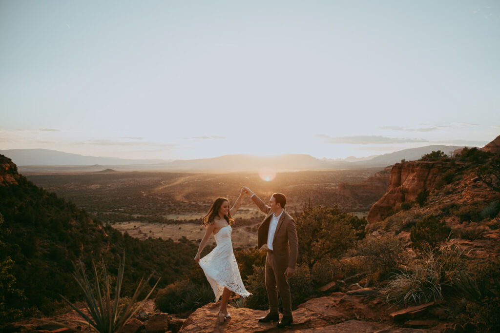 A man twirls a woman wearing a white dress while posing for Sedona engagement photos on Doe Mountain