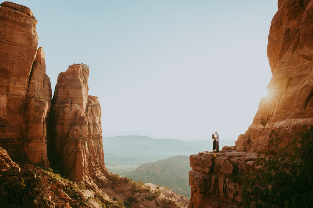 A couple stands at the edge of a cliff holding each other beneath a blue sky and near red rocks in Sedona. 