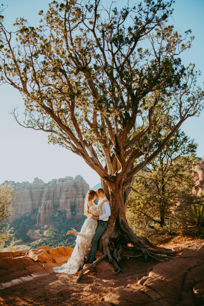 A man and woman lean in for a kiss against a tree as she kicks out her high heel at Merry Go Round Rock for their Sedona engagement photos