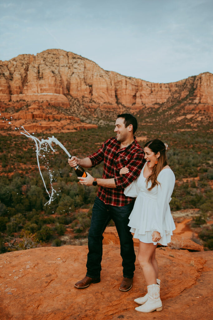 A man wearing a plan flannel shoots off a bottle of champagne at Courthouse Vista with his wife smiling and laughing while holding his arm for their Sedona engagement photos 