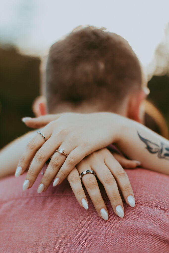 A detail shot of a woman's hands wrapped around her husband's shoulders with her rings and tattoo on her arm during a Sedona engagement photo shoot.
