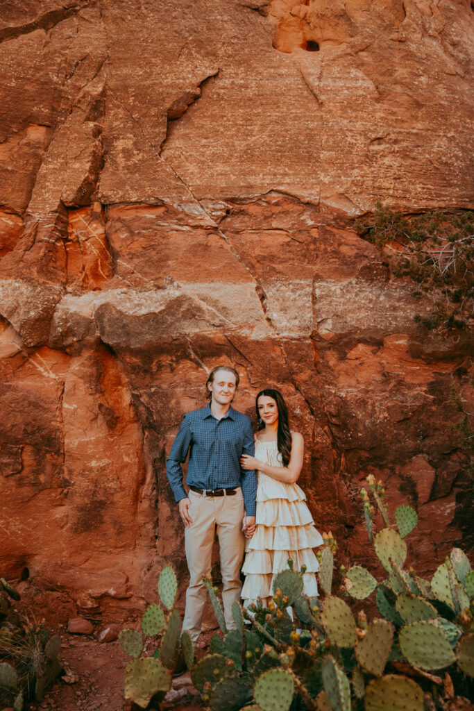 A couple stands side-by-side as they stand behind cactus and in front of Sedona's red rocks at Cathedral Rock for their Sedona engagement photos.