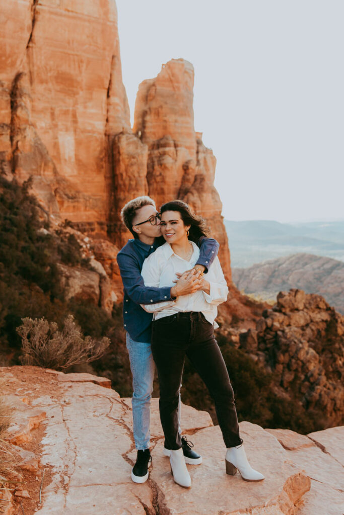 a woman kisses another woman's face as she holds her from behind for their Cathedral Rock engagement photos