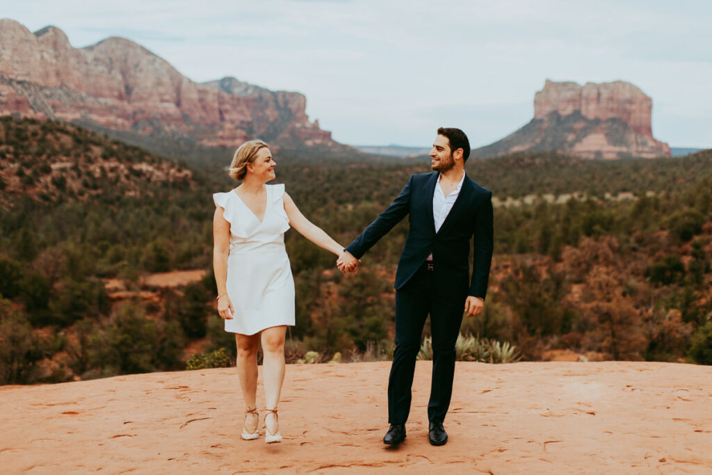 a couple wearing a classy outfit holds hands and looks at each other in front of a canyon with desert-green foliage and the red rocks of Sedona