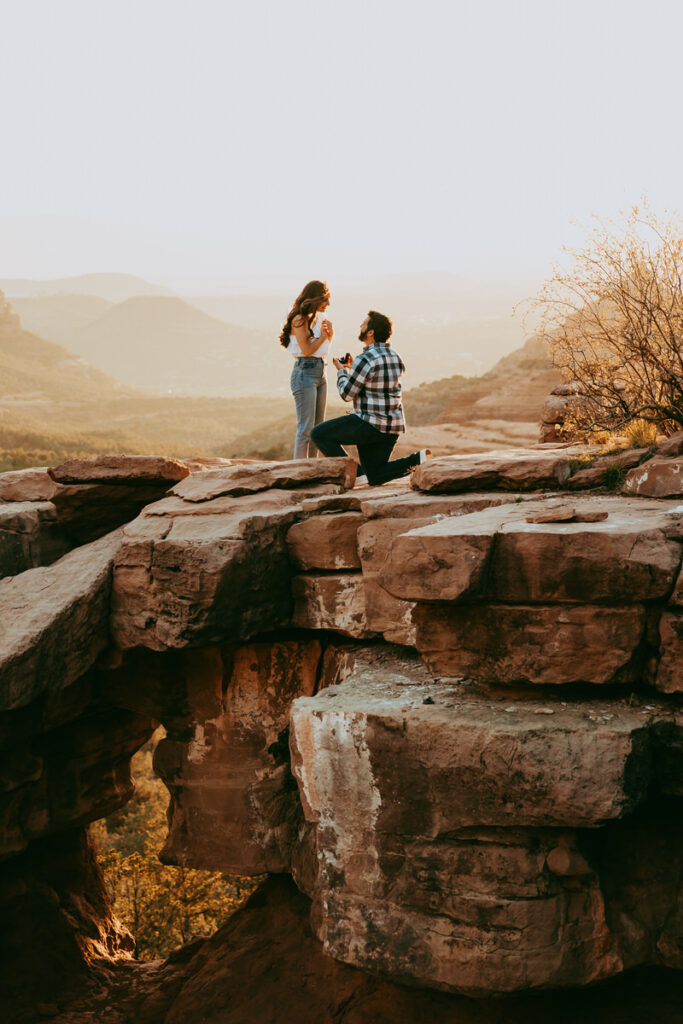 a man wearing a flannel is down on one knee proposing to a woman wearing casual jeans and a beautiful blouse in front of the beautiful canyons of Sedona