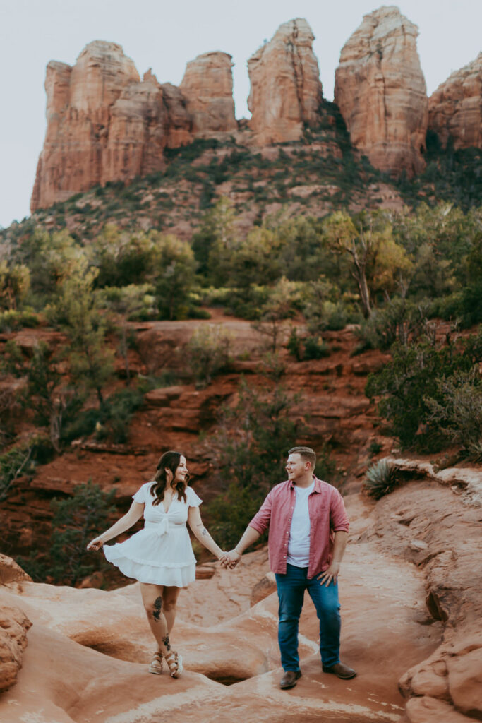 a woman with tattoos stands with one foot in front of the other wearing a cute white dress and holds hands with a man wearing a salmon colored overshirt, white shirt and blue jeans in sedona engagement photos