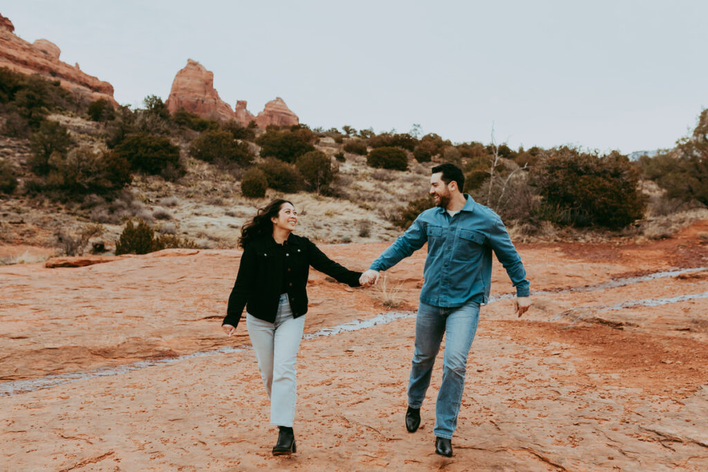 a man and woman smile as the run wearing multiple layers of clothes for their sedona engagement photos