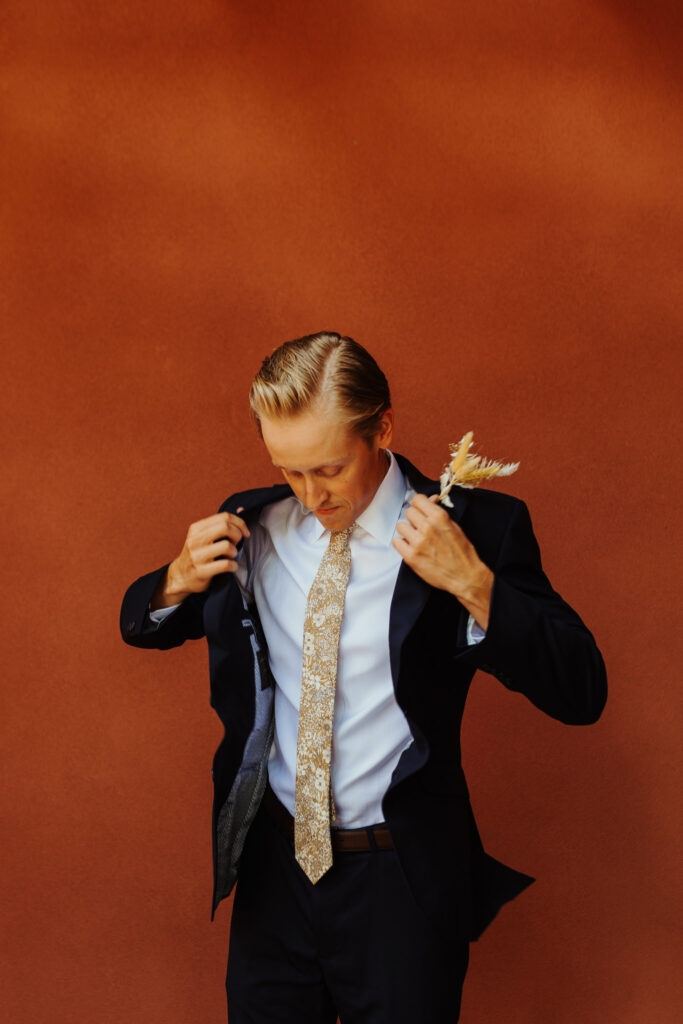man adjusts his wedding suit blazer while wearing it behind a striking red-orange painted wall in the Southwest