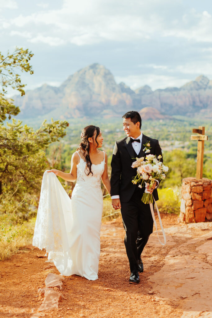 ride and groom hold each other's hands as they look at each other and walk toward camera with sedona red rocks in the background