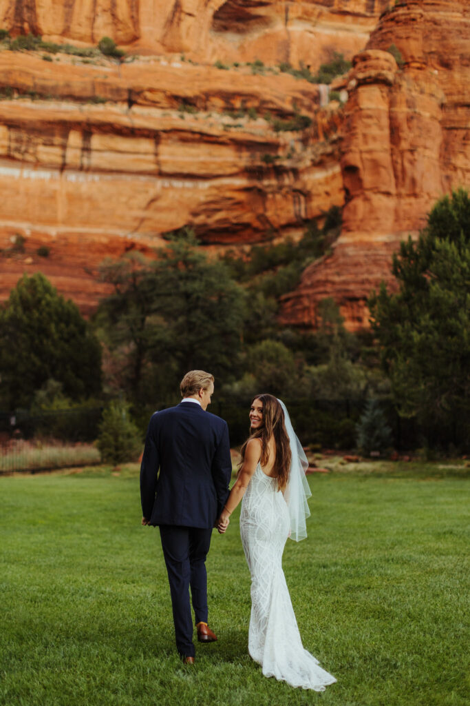 bride and groom hold hands as they walk away from the camera on green grass with tall sedona red rock background as beautiful bride walks back and looks at camera