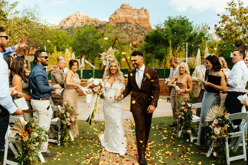 bride and groom smile as they walk down the aisle and guests throw flower petals in the air at amara resort with sedona red rocks in the backdrop